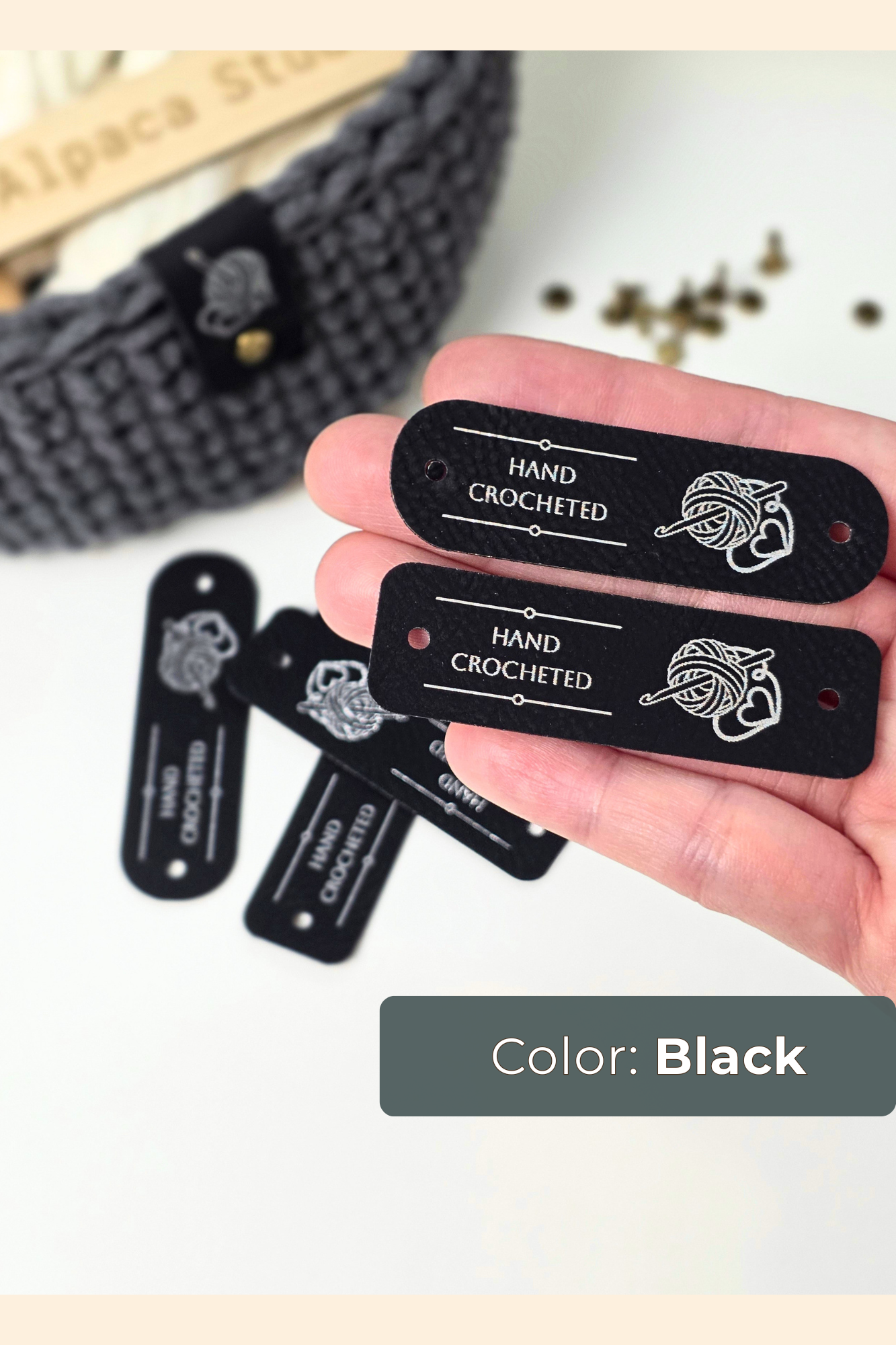 A hand holding two black rectangular labels printed with 'HAND CROCHETED' and a crochet hook graphic, with additional black labels scattered below. A crocheted gray item and a beige surface are blurred in the background, and a label at the bottom reads 'Color: Black'.