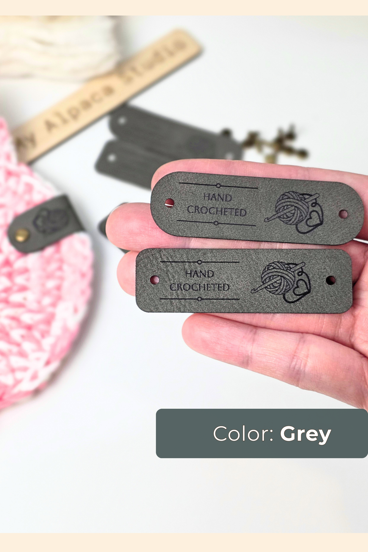 A hand holding two grey rectangular vegan leather tags engraved with 'HAND CROCHETED' and a small yarn logo. The background is a white surface with soft, blurred craft supplies, and a label at the bottom reads 'Color: Grey'.