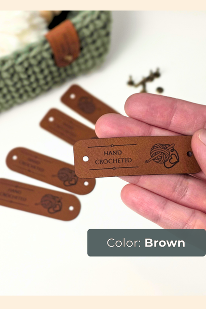 A hand holding a brown vegan leather tag engraved with 'HAND CROCHETED' and a yarn motif, with several matching tags scattered in the background. A dark gray label at the bottom reads 'Color: Brown'.