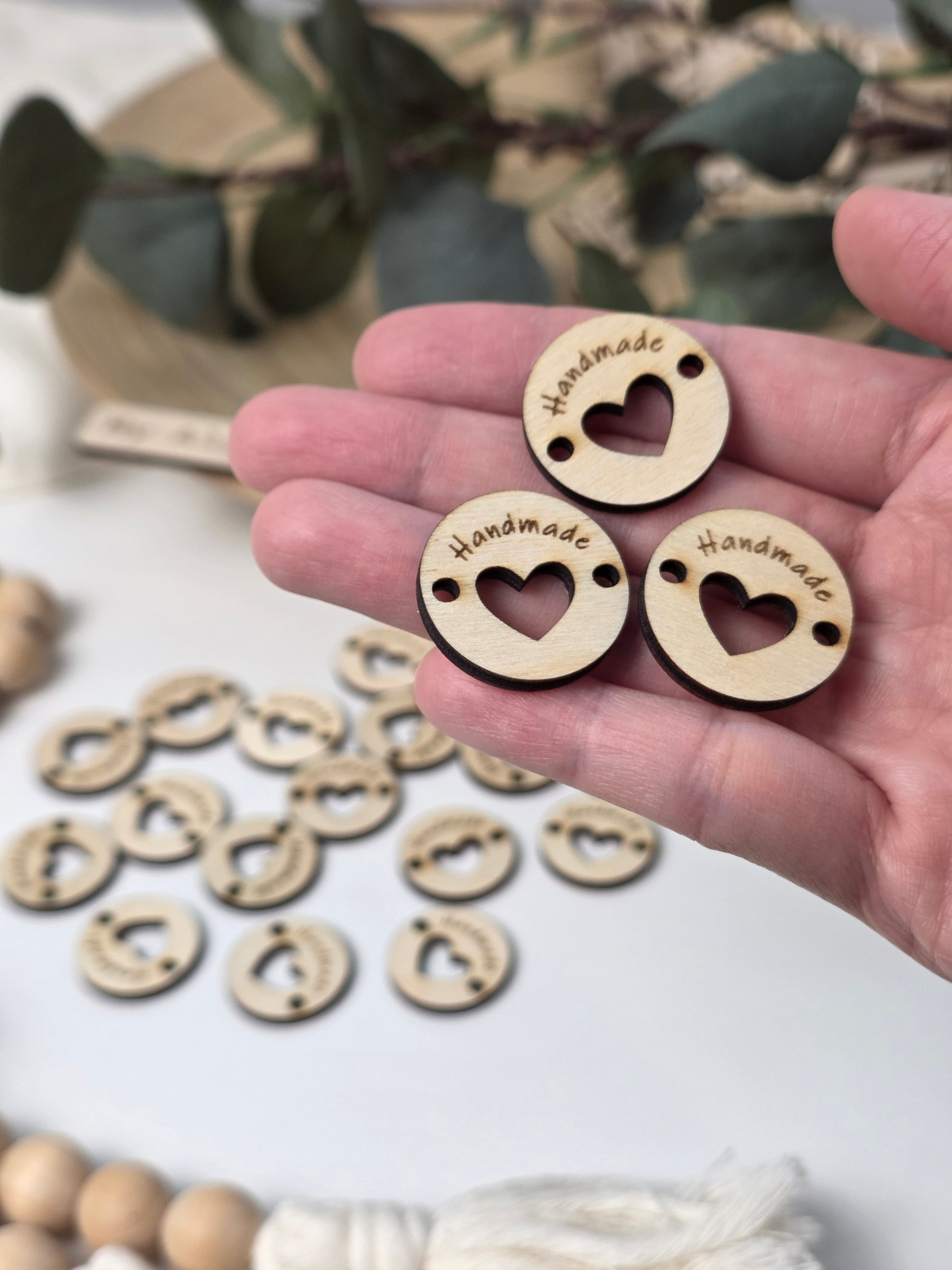 A hand holding three small round wooden beads engraved with 'Handmade' and a heart cutout, with more similar beads scattered in the background on a white surface.