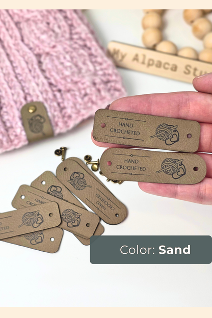 A hand holds several small rectangular sand-colored leather tags engraved with 'HAND CROCHETED' and a yarn ball motif, with more tags scattered below. A pink knitted textile and a wooden bead sign reading 'My Alpaca Studio' appear in the background, and a label at the bottom reads 'Color: Sand'.