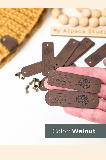 A hand holds several dark brown veagn leather tags engraved with 'HAND CROCHETED' and a logo, with brass eyelets beside them. The background is a light studio surface with a crocheted textile and beads, and a label at the bottom reads 'Color: Walnut'.