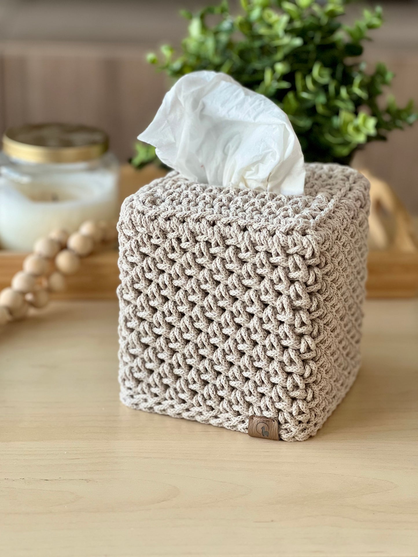 Knitted tissue box cover with tissues on a wooden surface, surrounded by decorative items.