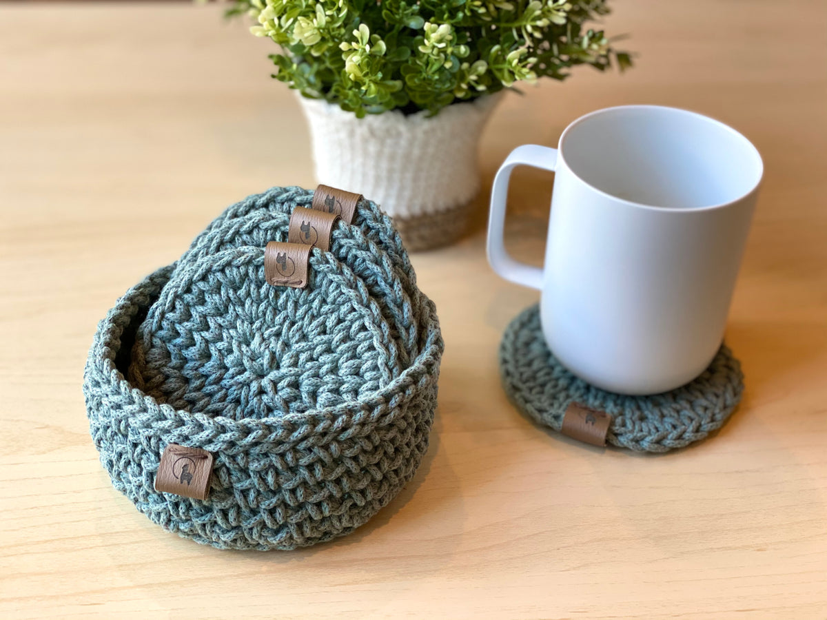 A crochet kit for making four coasters and one holder, displayed with yarn, a crochet hook, and a pattern booklet.