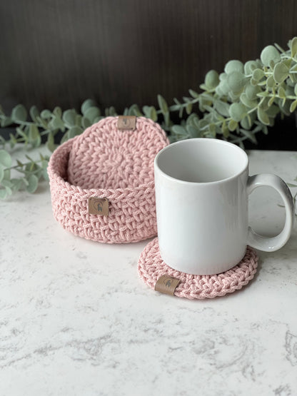 A white ceramic mug sits on a round pink crocheted coaster, with a matching pink crocheted basket behind it on a light marble surface and green foliage in the background.